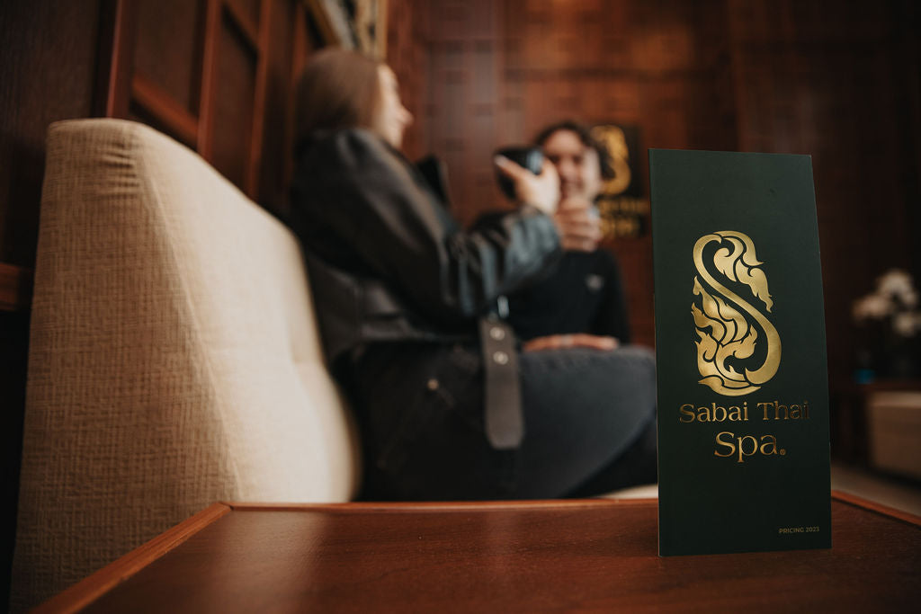 Sabai Thai Spa brochure on a wooden table with blurred background of two people conversing on a sofa.