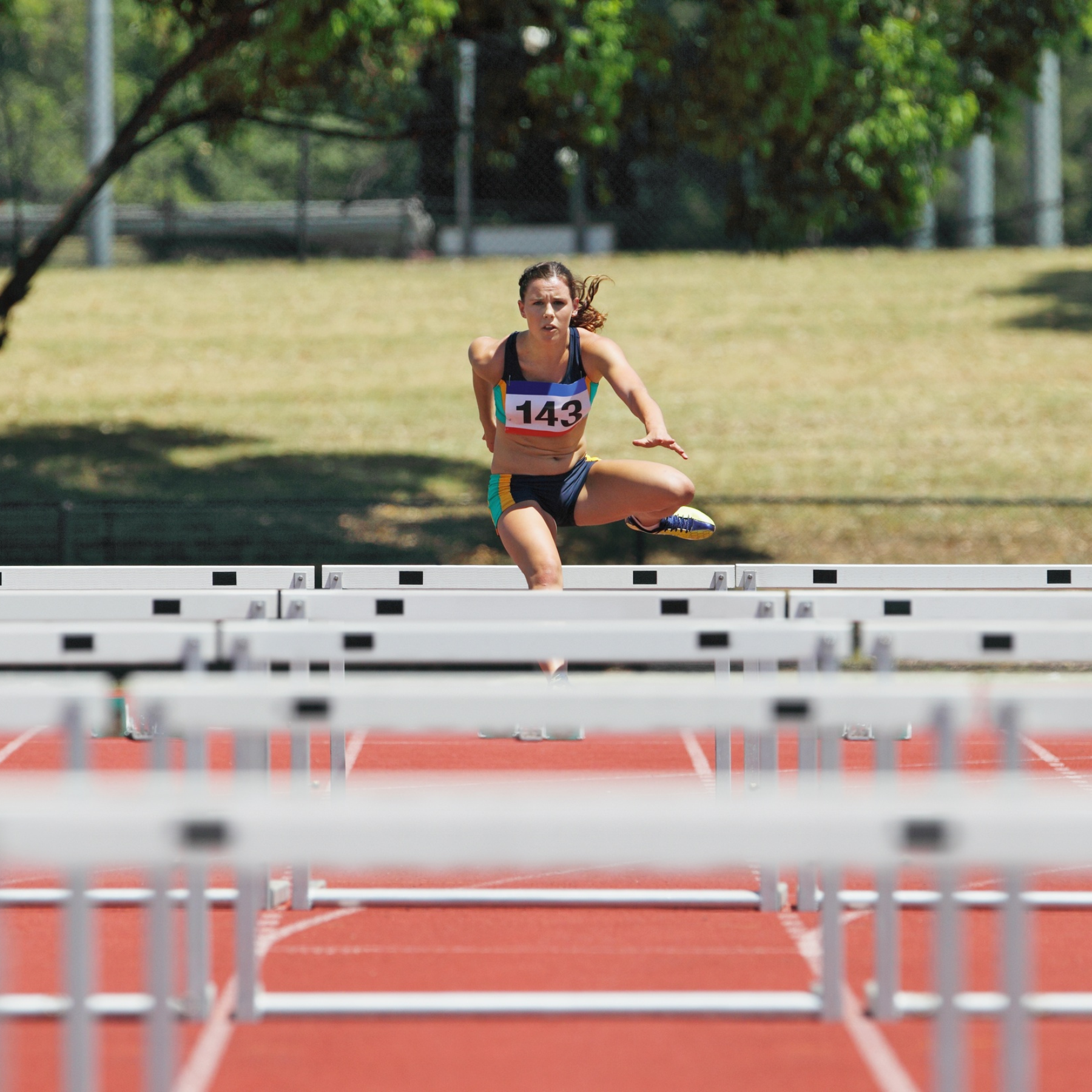 A female athlete wearing a race bib with the number 143 is mid-air while jumping over a hurdle on a red running track, displaying focus and determination.