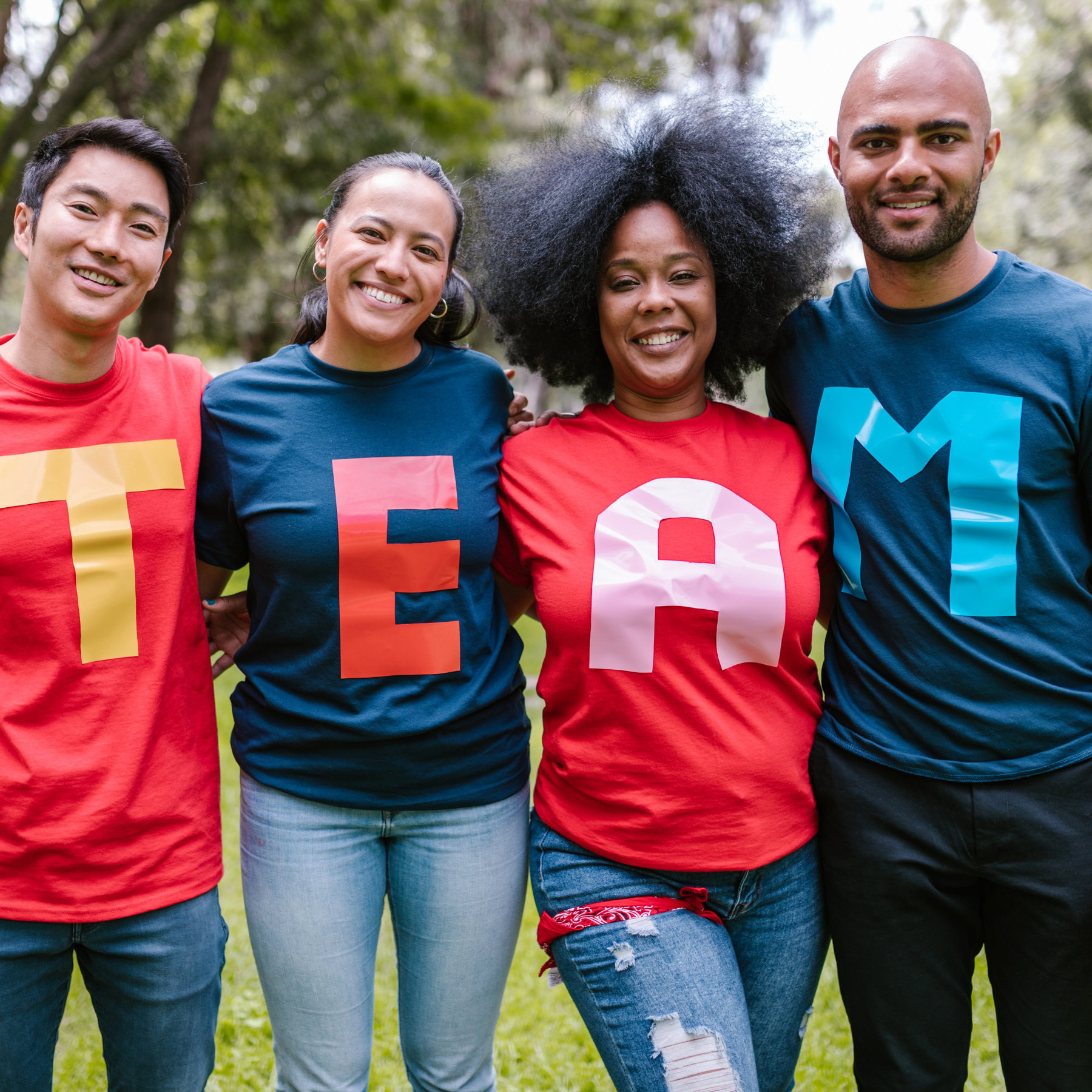 Four people standing arm-in-arm in a park, wearing colorful T-shirts with large letters spelling 'TEAM.' They are smiling with trees and grass in the background.