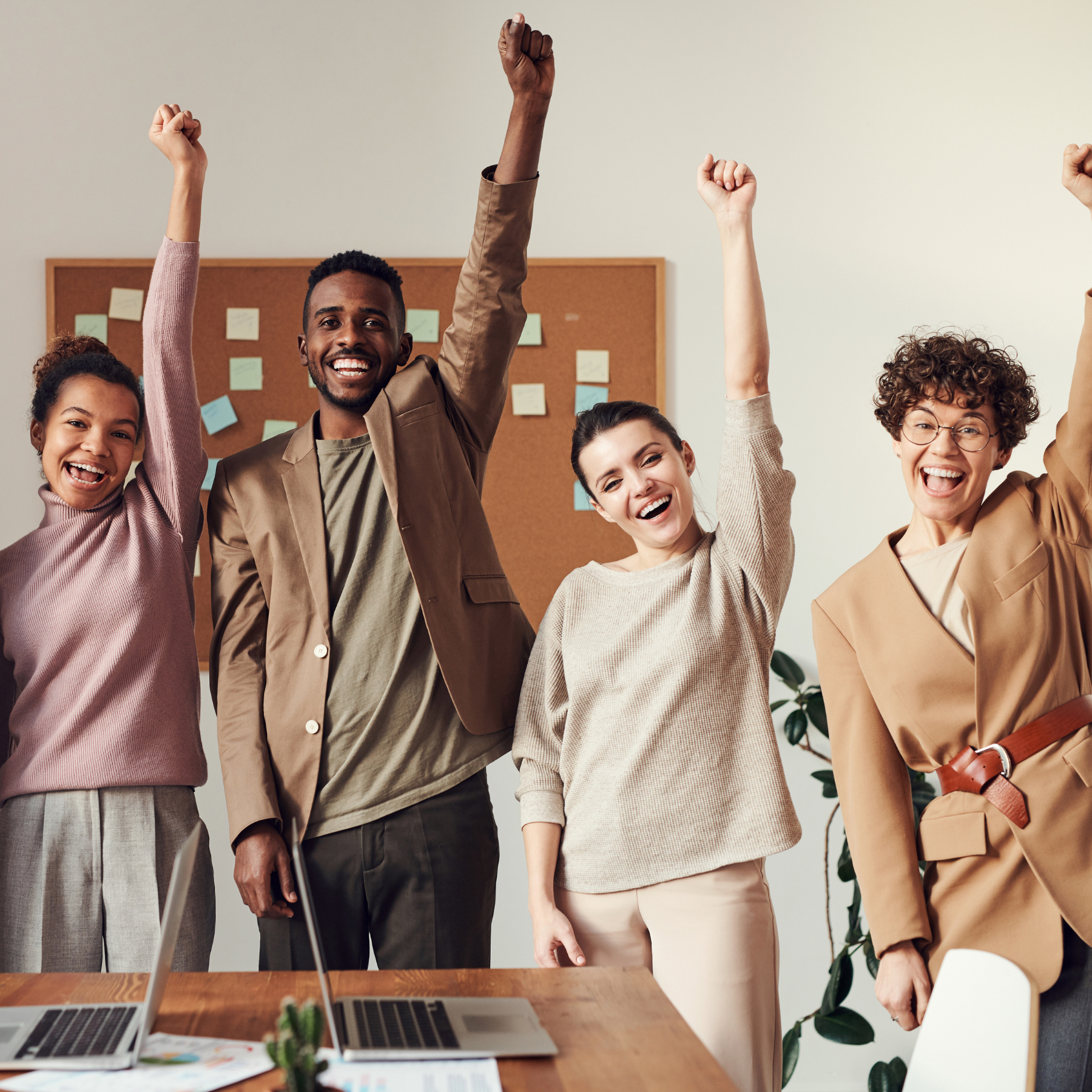 Four cheerful colleagues standing with raised arms, smiling in celebration in a bright office. A corkboard covered with sticky notes is visible in the background.