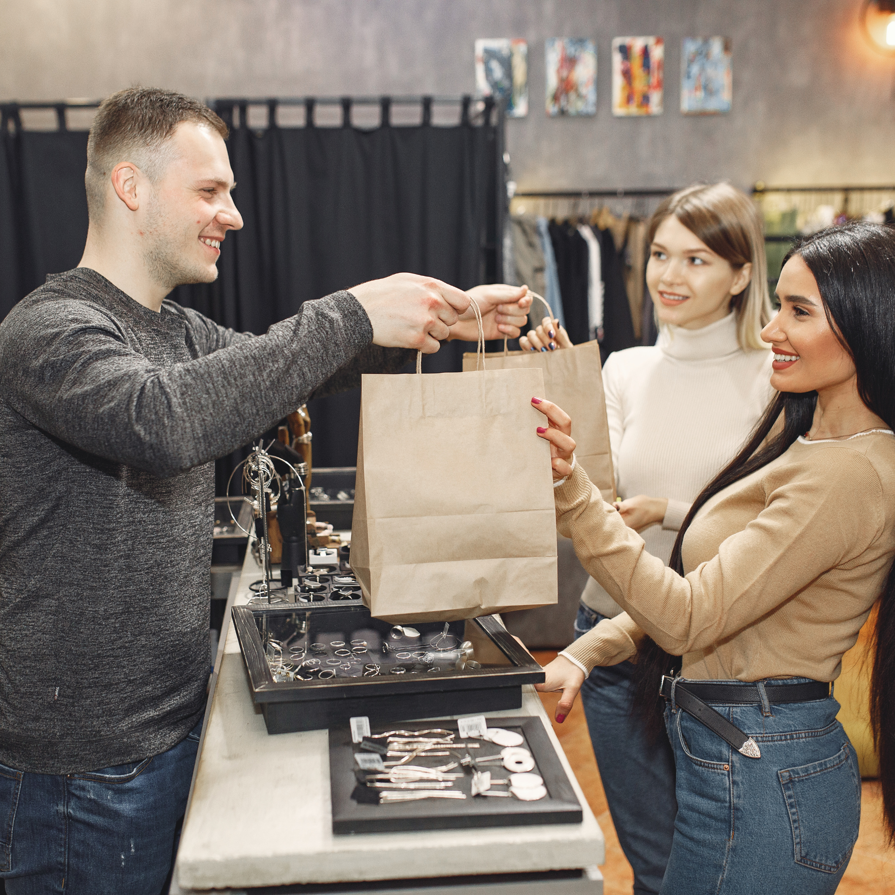 A smiling shopkeeper hands a paper bag to a customer while another woman watches, inside a jewelry store.