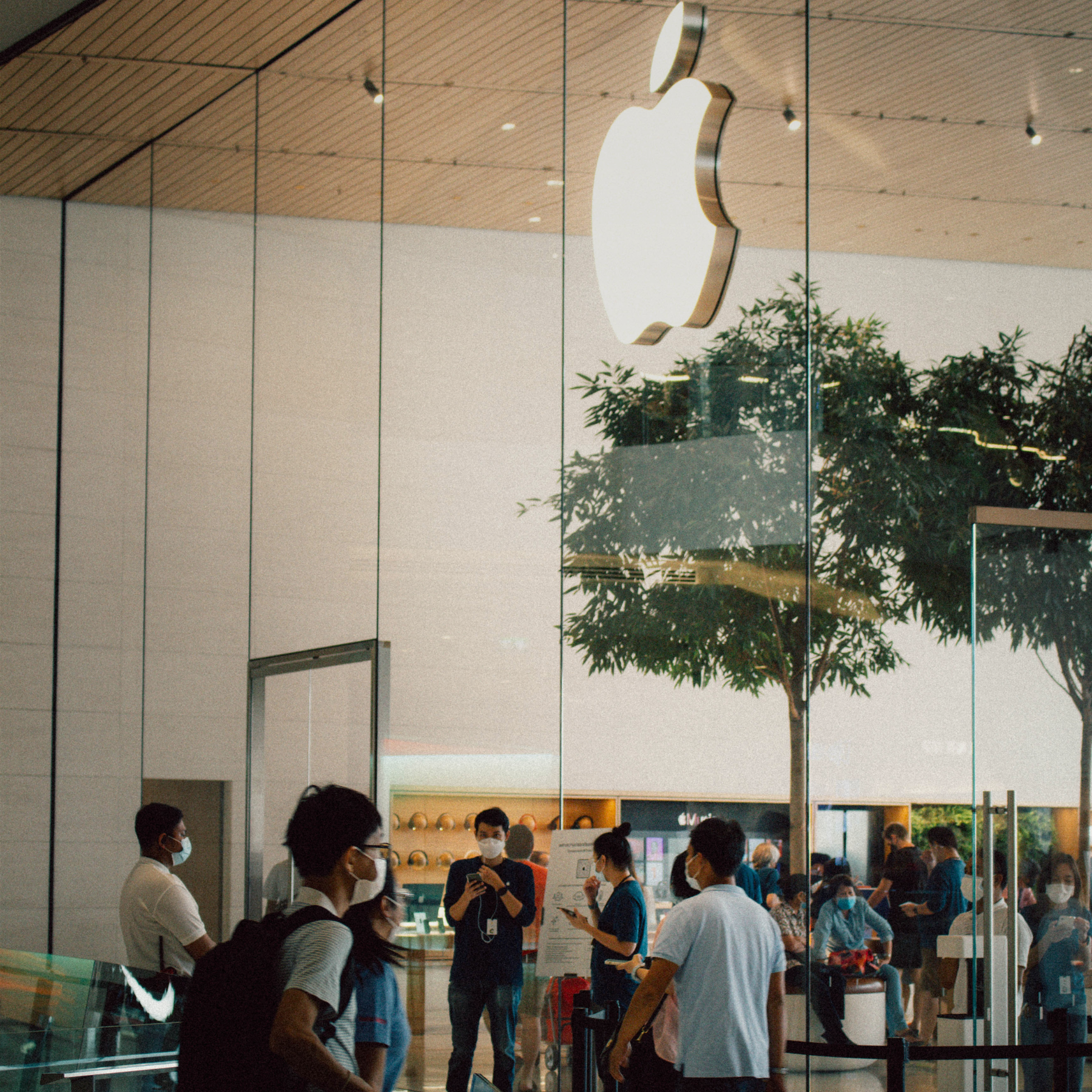 A busy Apple Store with a glass facade, large Apple logo, and indoor trees. Customers and employees, many in face masks, interact inside.