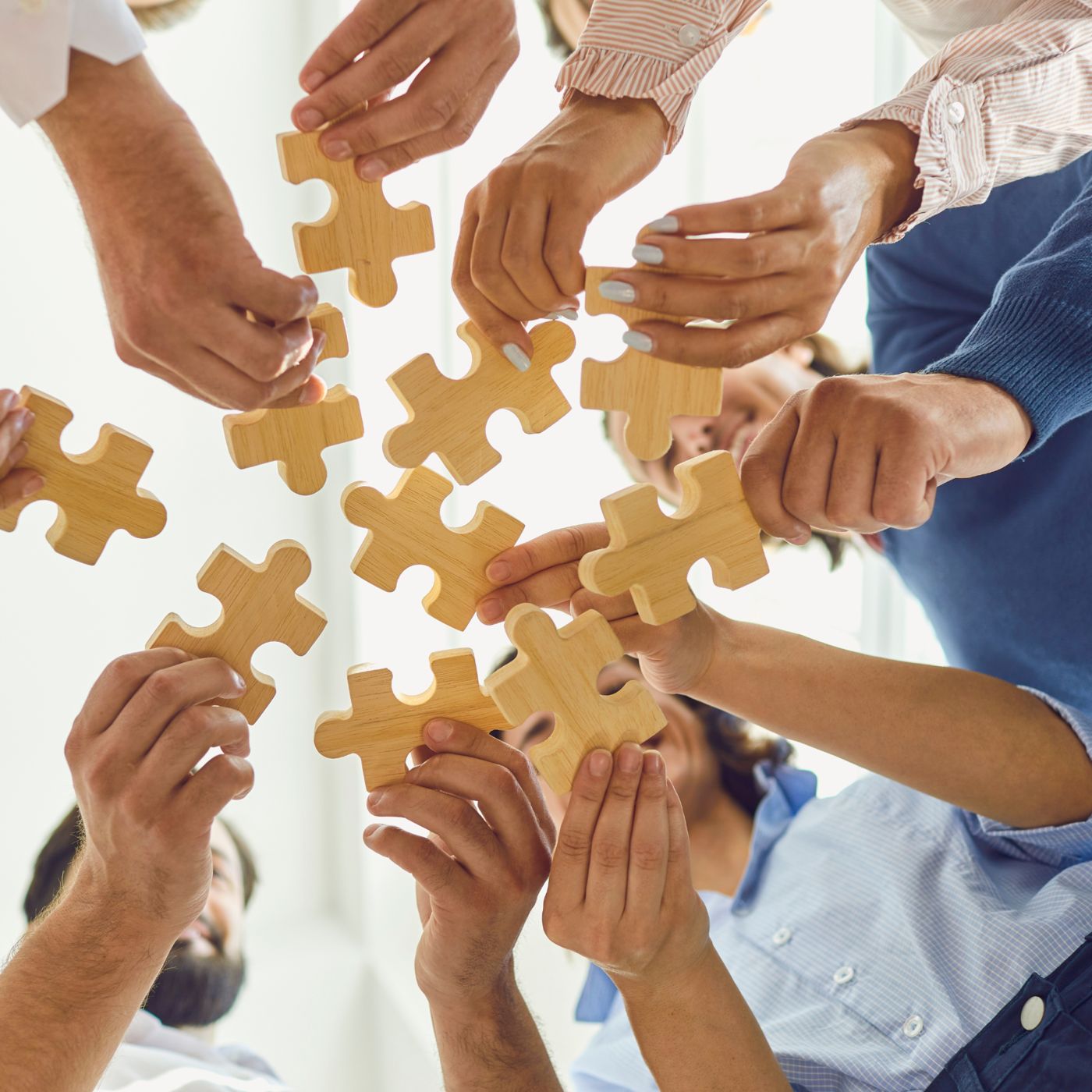 A group of people holding wooden puzzle pieces, reaching their hands toward the center to connect them, with a bright background.