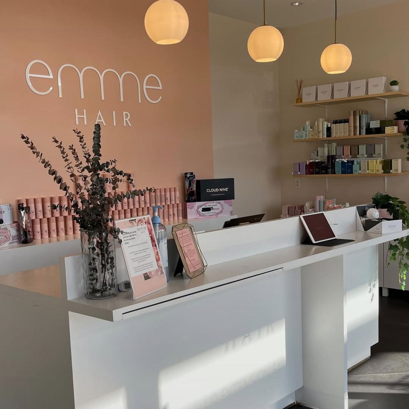 Reception area of Emme Hair Vancouver salon with modern white counter, branded wall sign, hair care products, and warm pendant lighting.