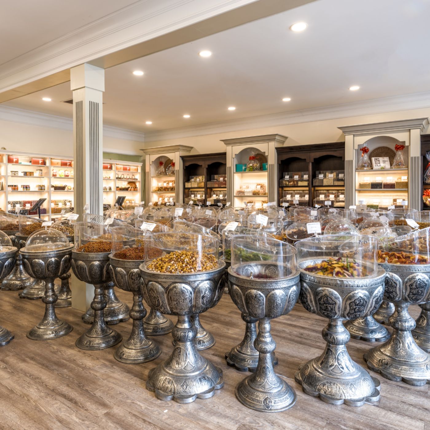 A specialty store filled with ornate, silver pedestal containers holding a variety of dried fruits, nuts, and snacks. Each container has a clear plastic cover with labels. The background features elegant wooden shelves stocked with packaged goods, softly lit by warm lighting. The store has a clean, inviting atmosphere with a blend of traditional and modern design elements.