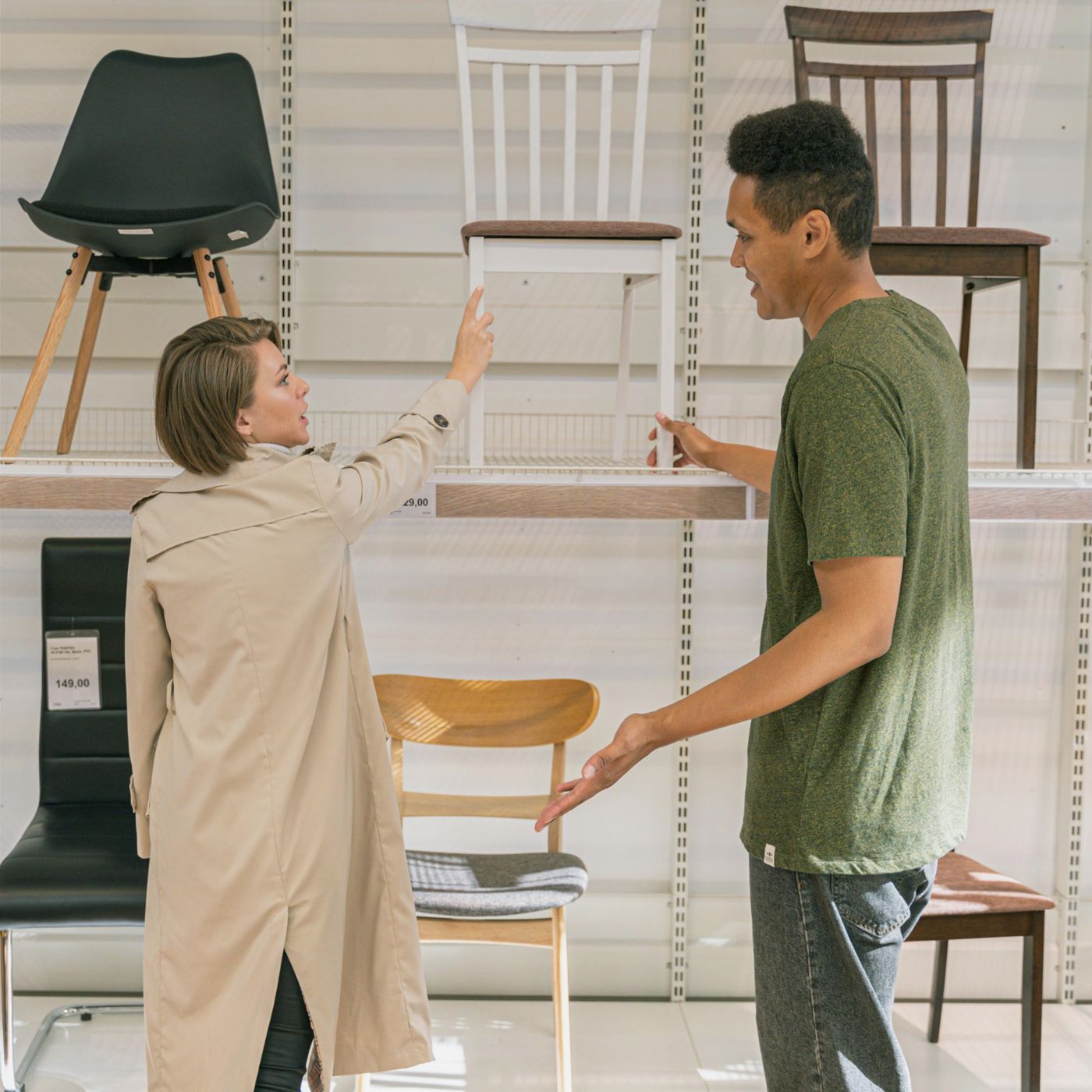A woman in a beige trench coat and a man in a green t-shirt are shopping for chairs in a furniture store. The woman points at a white chair on a shelf while the man gestures toward it, seemingly discussing their choice. Various chairs in different styles and colors are displayed on shelves behind them. The store is well-lit with a modern, minimalistic design.