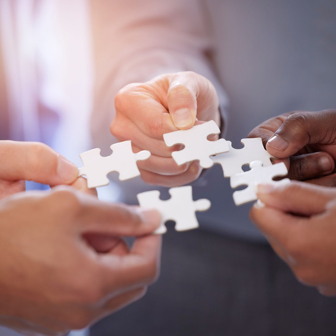 Four hands, each holding a white puzzle piece, coming together to form a complete connection, symbolizing teamwork and collaboration.