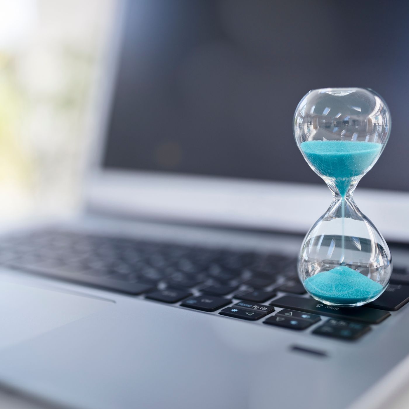A small glass hourglass with blue sand placed on a laptop keyboard, symbolizing time management and deadlines.