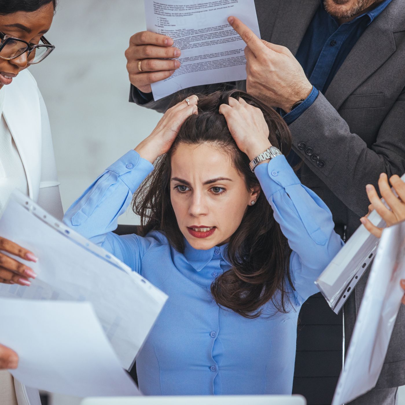 A stressed woman in a blue shirt sitting at her desk, holding her head in frustration as colleagues surround her with documents and demands.