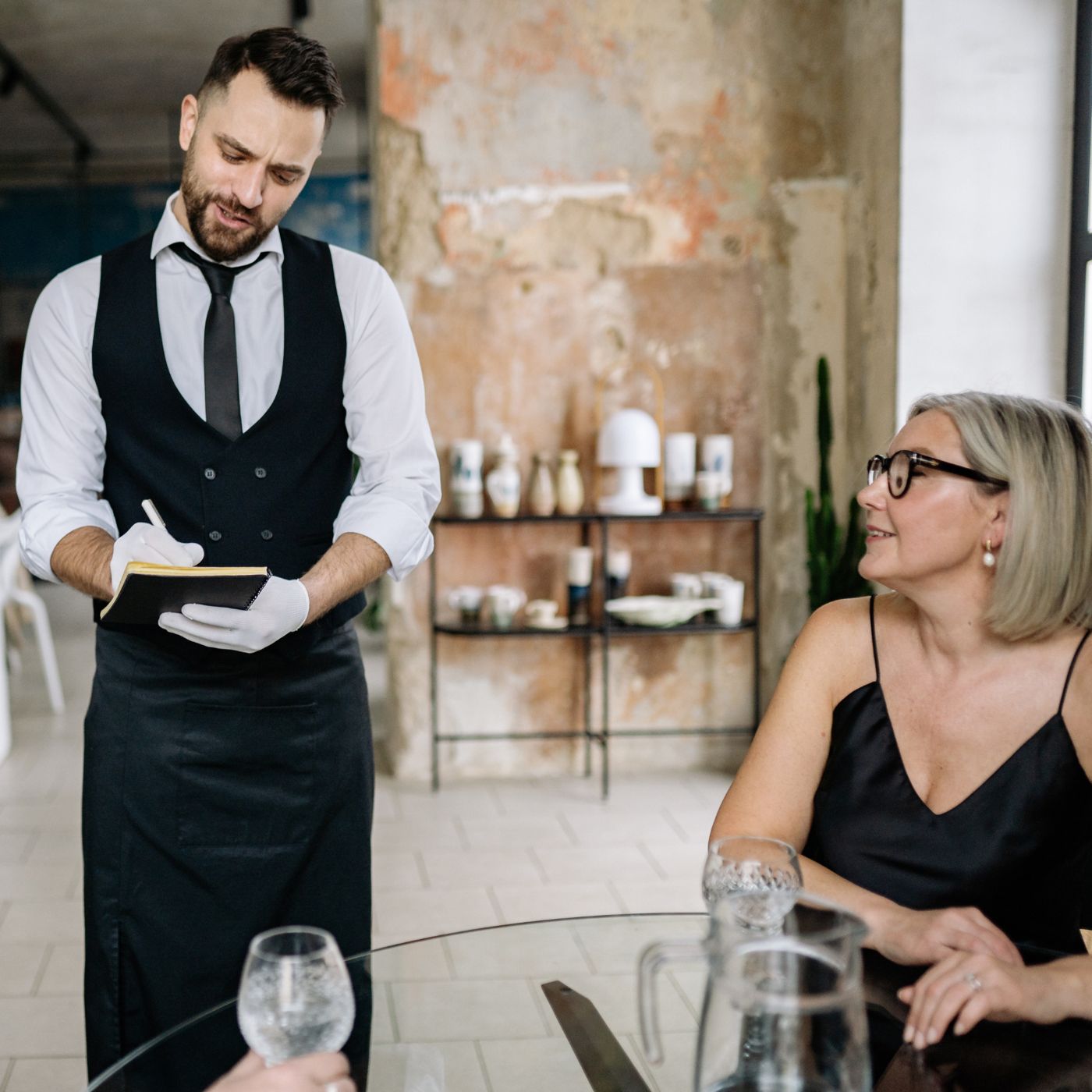 A smiling waiter in a black vest and gloves takes an order from a woman in a black dress at a stylish restaurant.