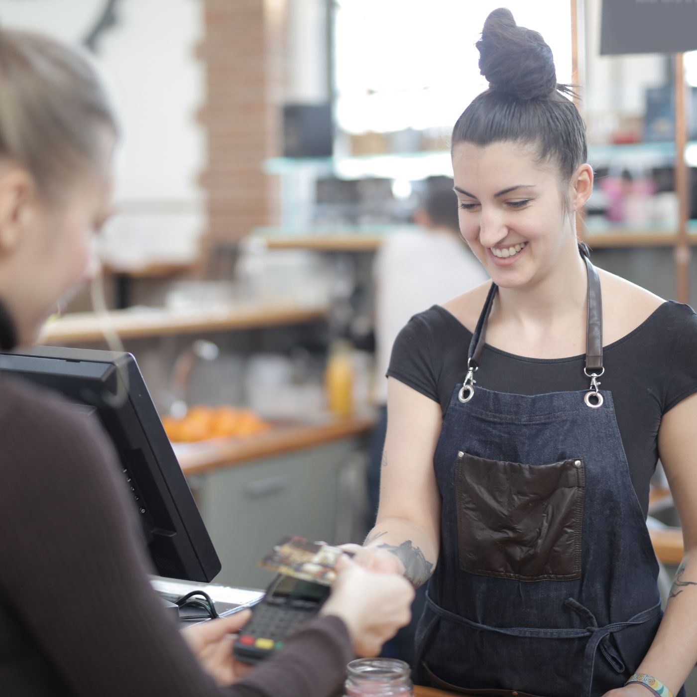A smiling barista in a denim apron hands a receipt to a customer paying with a card at a coffee shop.
