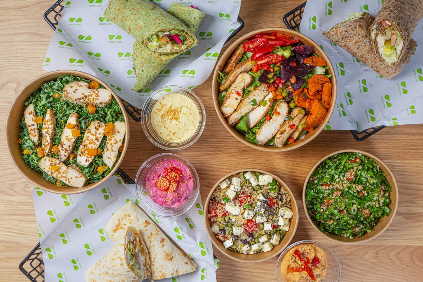 Overhead view of colorful bowls, wraps, and dips arranged neatly on a table, featuring fresh salads and chicken dishes.
